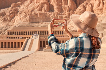 Travel blogger girl takes pictures on a smartphone at the famous Hatshepsut temple in the ancient city of Luxor in Egypt. - obrazy, fototapety, plakaty