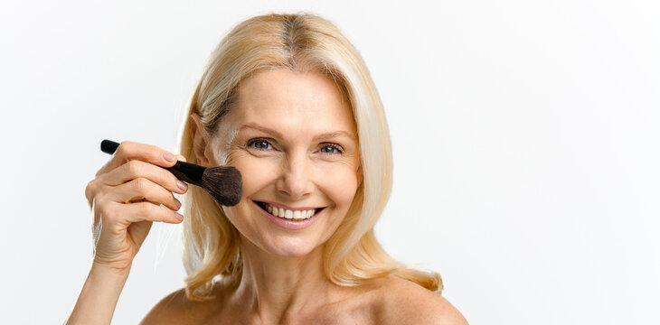 Charming blonde 40s 50s middle aged woman with holding make up brush on shining face with perfect skin, looking at the camera, applying powder or some cosmetic product, advertising concept