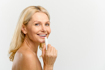 Happy and optimistic beautiful middle age woman applying natural lip balm isolated on white. Charming blonde lady using moisturizing lipstick, treatment balm. Advertising concept