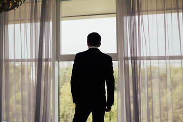 The man in black suit standing near the panoramic window with the green trees background. - Powered by Adobe