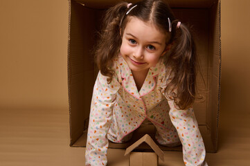 Cute child girl peeking out from behind a cardboard box, looking at camera. Craft carton box on a...