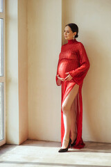 Portrait of attractive pregnant woman in beautiful shiny red dress standing near wall in photostudio, holding her pregnant belly and looking out the window