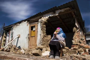 Fotobehang War in Ukraine. Grandmother prays for an end to the war. Ukrainian refugees. A woman in front of a house destroyed by a rocket. Consequences of the war in Ukraine. Ukrainian woman prays for peace. © Pavlo