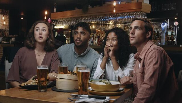 A multi-ethnic group of friends watching a sports game on TV at a restaurant. They cheer and high five each other when their team scores.