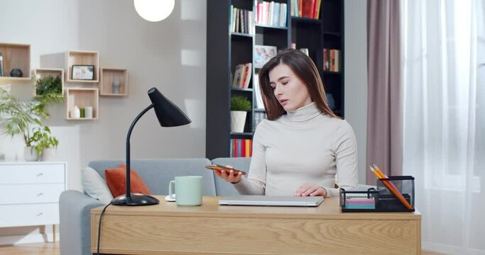 Beautiful Caucasian woman closing laptop and taking smartphone in hands. At home. Pretty young female freelancer sitting at desk in cozy room and finishing work at computre. Scrolling on phone.