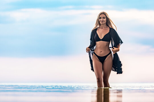 A girl with blond chic hair in a black bikini and a lace shawl walks along the estuary