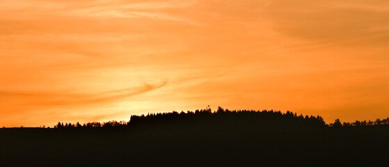 Orange clouds on the horizon at sunset over a hill covered with forest.