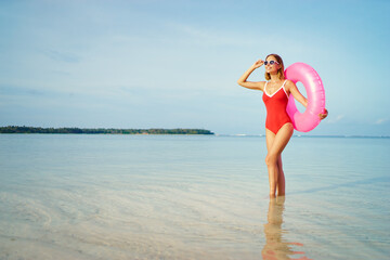 Naklejka na ściany i meble Enjoying suntan and vacation. Outdoor portrait of pretty young woman in red swimsuit with inflatable ring on tropical beach.