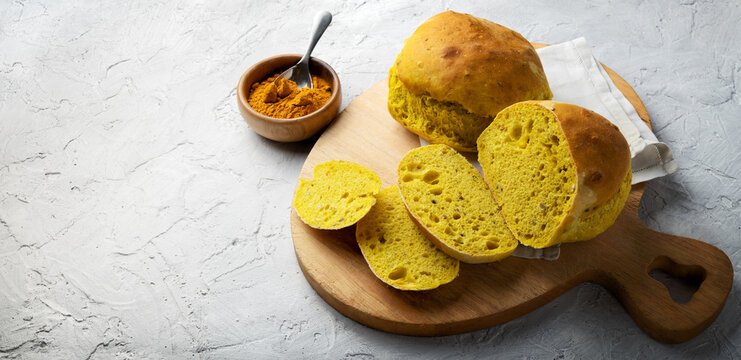 Turmeric bread, two tumeric bread rolls, one cut into slices on wooden cutting board. Bowl with turmeric powder on white plaster background. Space for text.