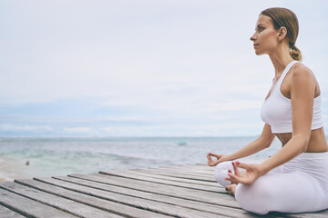 Fototapeta na wymiar Yoga and meditation. Closeup of young woman in lotus pose on wooden deck with sea view.
