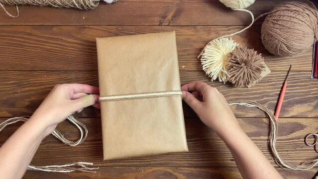 Woman ties gift box in craft paper with twine. Yarn pom-poms on wooden table. Top view of festive composition.
