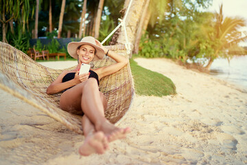 Fototapeta premium Vacation and technology. Young pretty woman in hat and swimsuit using smartphone laying in hammock on the beach.