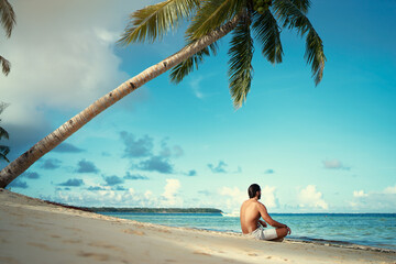 Suntan and meditation. Enjoying vacation.. Relaxed young man siiting on lotus position on the tropical beach.