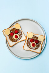 Funny bear face sandwich toast bread with peanut butter, cheese and raspberry on plate blue background. Kids child sweet dessert breakfast lunch food close up