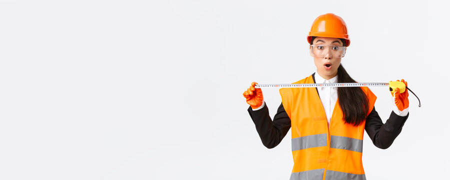 Surprised And Excited Asian Female Construction Engineer, Industrial Worker In Safety Helmet And Uniform, Measuring Layout, Holding Tape Measure And Stare Amazed At Camera, White Background