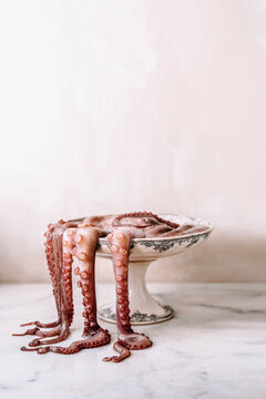 An octopus in an old and decorated footed dish, the tentacles falling outside on a marble table.