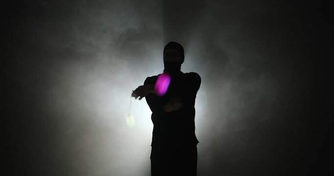 Silhouette of young man in balaclava mask spinning led poi hand props in illuminated background with smoke with smoke