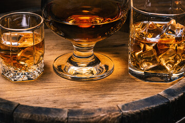 Alcoholic beverages whiskey, cognac, brandy, stand in glasses with ice on wooden barrel of whiskey close-up. Drink whiskey or cognac with ice in bar and spilled drinks directly from barrel.
