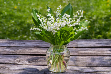 Lily of valley flower in vase on wooden table on summer day in rays of sunlight close-up. Lilies of valley are forested against green background.