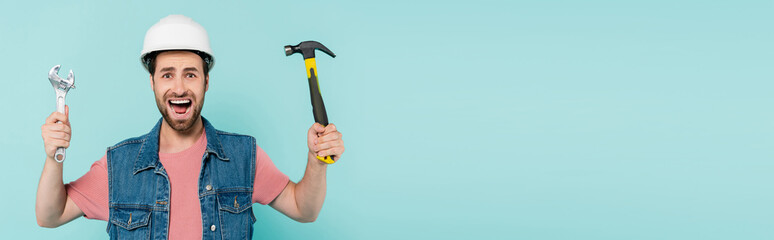 Screaming man in helmet holding hammer and wrench isolated on blue, banner.