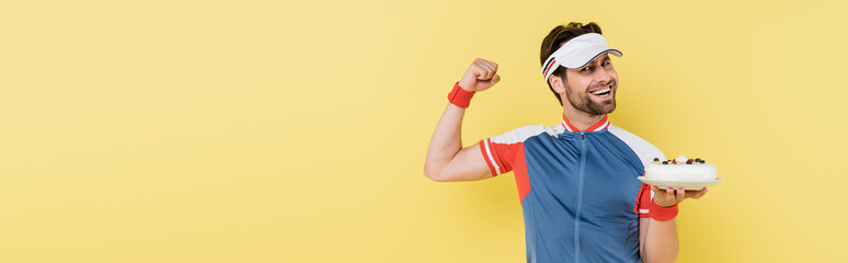 Cheerful sportsman holding cake and showing muscles isolated on yellow, banner.