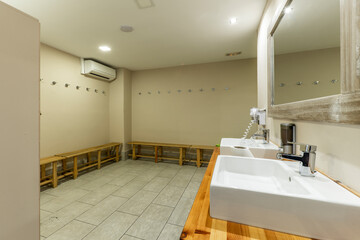 White porcelain sinks under a mirror in the common bathrooms of a gym with wooden benches around the perimeter