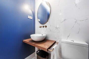 Charming bathroom with an oval mirror, a hemispherical porcelain sink on a solid wood countertop, painted blue walls and floors, and another wall covered in white marble with gray veins