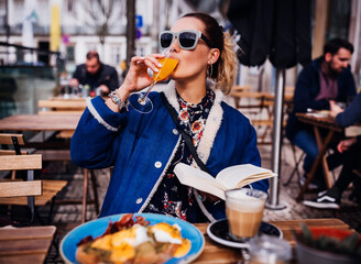 Lifestyle fashion portrait of pretty girl having brunch in outdoors cafe. Drinking an aperitif and enjoying meal. Reading book, enjoying vacation. Wearing stylish jeans coat, sunglasses. Holiday