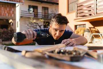 A man is using a screwdriver drill on wood on his workbench in his backyard during the day