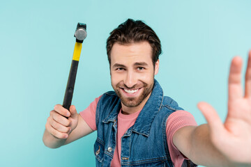 Smiling man looking at camera while holding hammer isolated on blue.