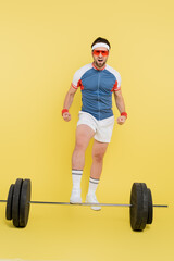 Fototapeta na wymiar Young sportsman in sunglasses showing muscles near barbell on yellow background.