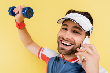 Smiling sportsman talking on smartphone and looking at camera while training with dumbbell isolated...