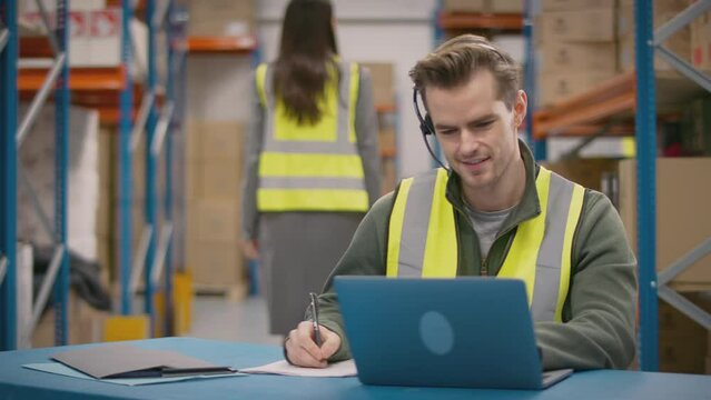 Male worker wearing headset and high vis vest sitting at laptop in busy distribution warehouse - shot in slow motion