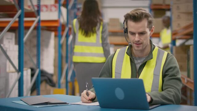 Male worker wearing headset and high vis vest sitting at laptop in busy distribution warehouse - shot in slow motion