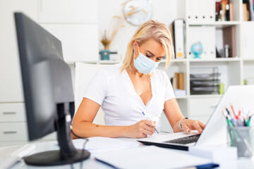 Woman in disposable face mask working in business office using laptop