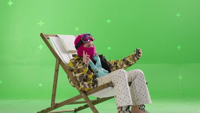 A young snowboarder sits in a sun lounger and takes a selfie against a green background in the studio. Active lifestyle in the mountains. Chroma key, tracking points