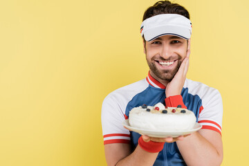 Positive sportsman in visor holding blurred cake isolated on yellow.