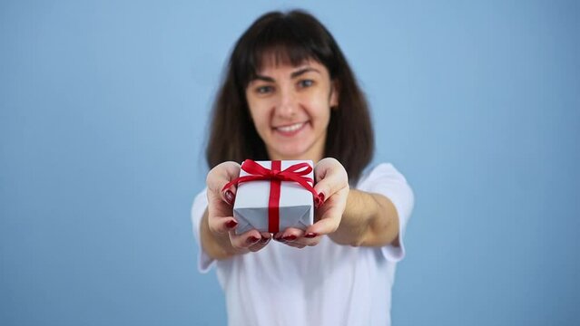 Smiling brunette woman holding gift box and gives it by hands to camera on blue wall background