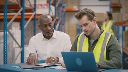 Manager with having meeting with male worker wearing headset and high vis vest sitting at laptop in busy distribution warehouse - shot in slow motion - Powered by Adobe