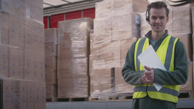 Male worker in freight haulage business wearing headset standing next to truck being loaded at distribution warehouse holding clipboard looking into camera - shot in slow motion