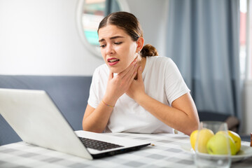 Young girl sitting at a table in front of a laptop in an apartment informs the doctor at the reception via video link that ..she has a sore throat