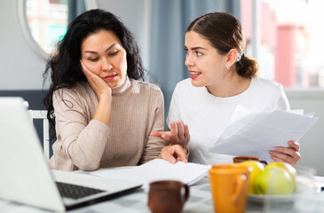 Two women with financial documents near laptop sitting at the table at home