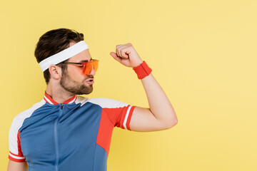 Brunette sportsman in sunglasses looking at muscles isolated on yellow.