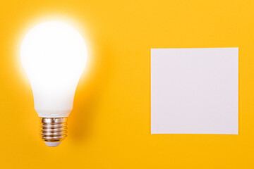 Glowing Power Saving Lamp with White Note Paper Lying on Yellow Table, Blank Template with Copy Space - Top View, Flat Lay
