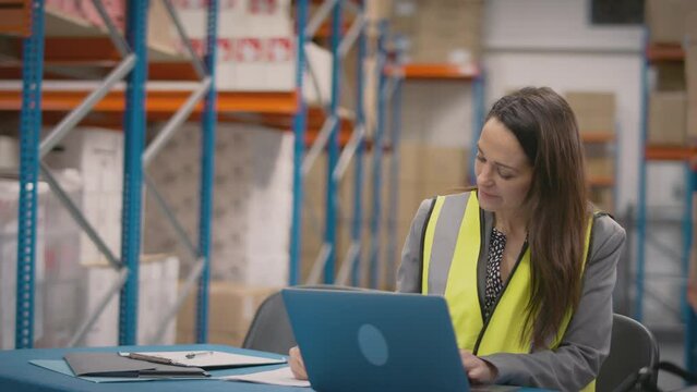 Female manager wearing high vis vest sitting at laptop in busy distribution warehouse - shot in slow motion