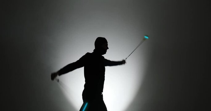 Silhouette of man in balaclava mask spinning led poi hand props in illuminated background with smoke