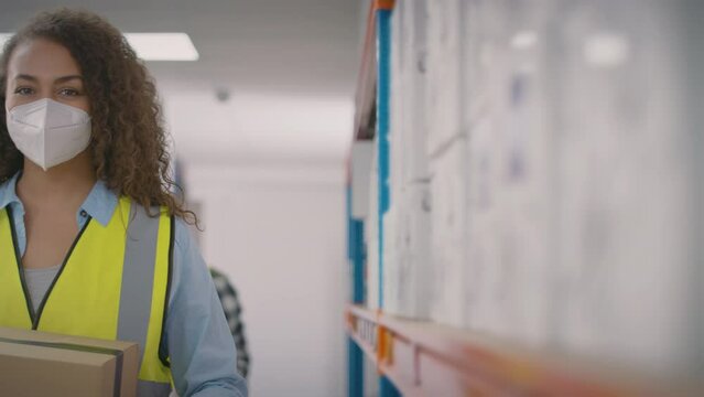 Camera tracks past warehouse shelves to show female worker in distribution warehouse wearing PPE face mask and looking into camera - shot in slow motion