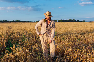 Naklejka na ściany i meble Rural Countryside. Tall handsome man dressed in a coarse linen suit and hat standing at golden oat field