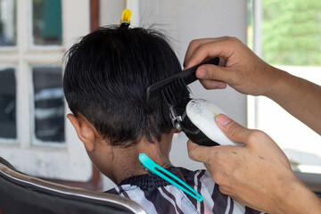 Hairdresser holding hair clipper for haircutting asian boy in barber shop.