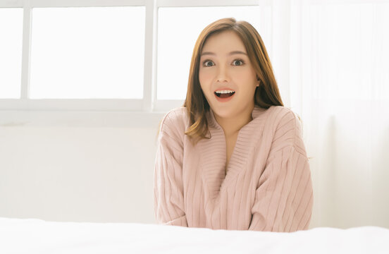 Portrait of an excited or surprised pretty Asian woman in warm knitted pink clothes feeling happy, astonished, lucky and surprised, looking at camera while sitting on floor in bedroom. Fashion, winter